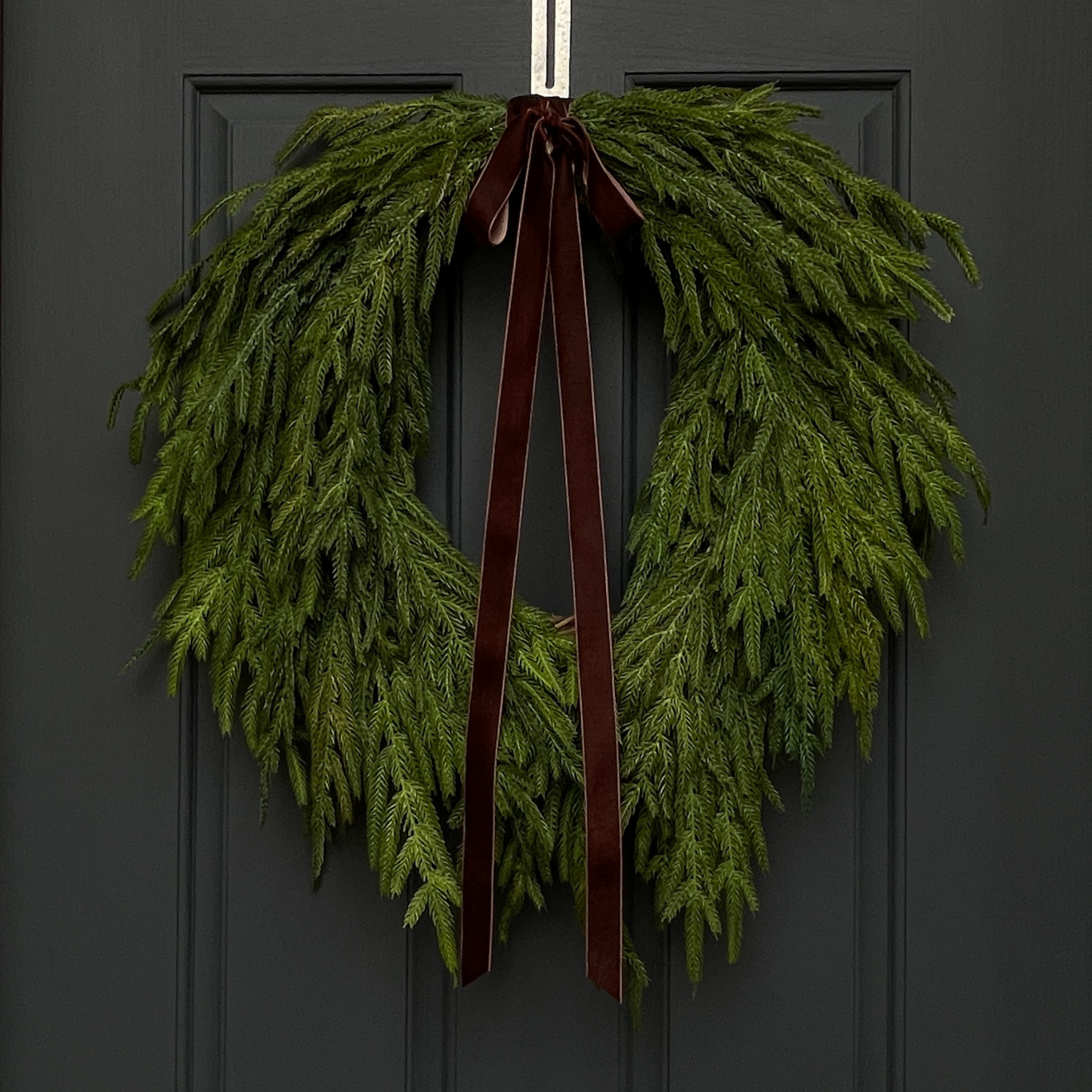 A faux Christmas greenery wreath with real touch norfolk pine and a dark brown velvet bow, hanging on a dark door.
