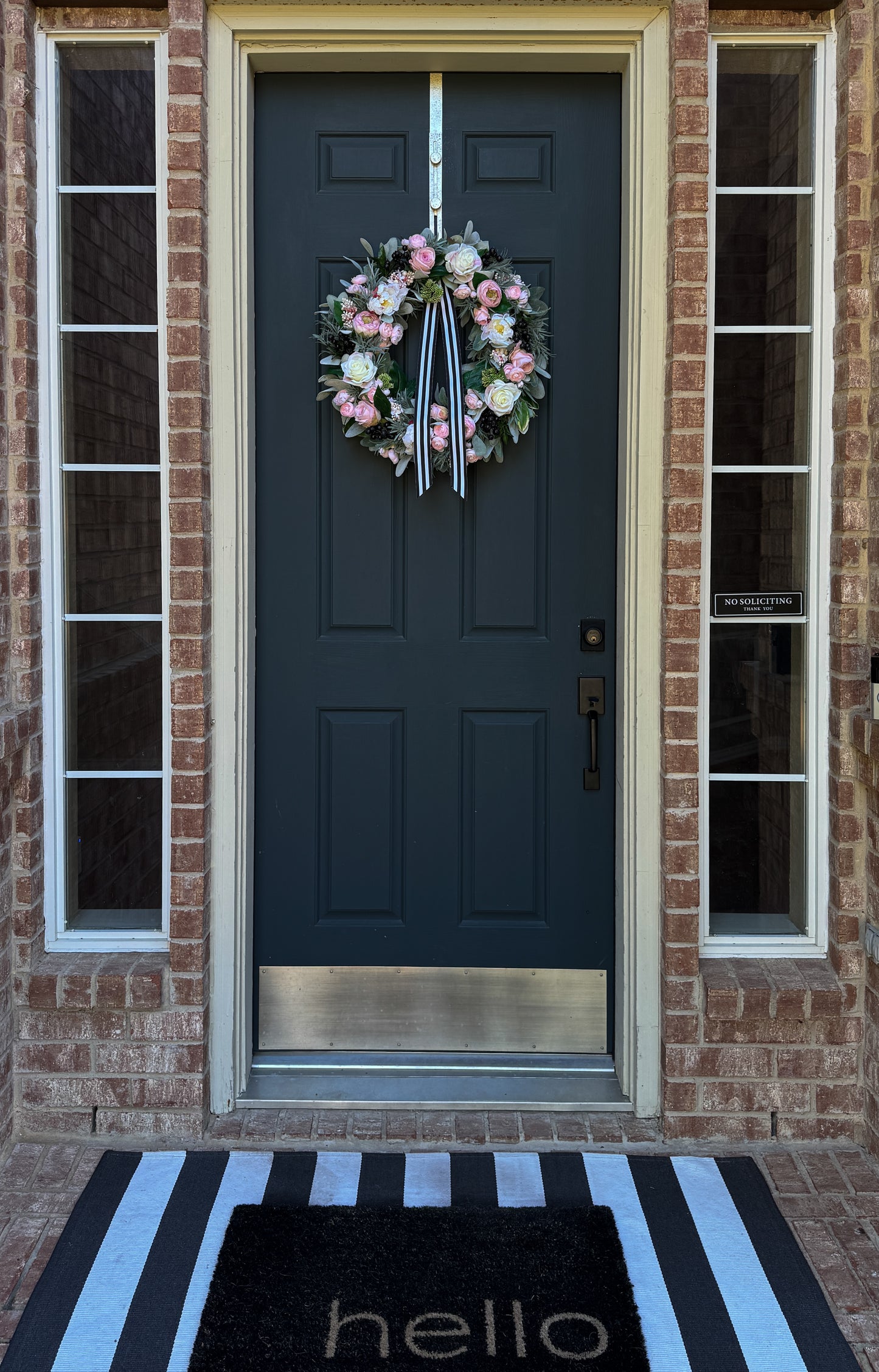 Front door with a floral wreath, striped doormat, and brick wall.
