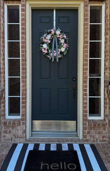 Front door with a floral wreath, striped doormat, and brick wall.