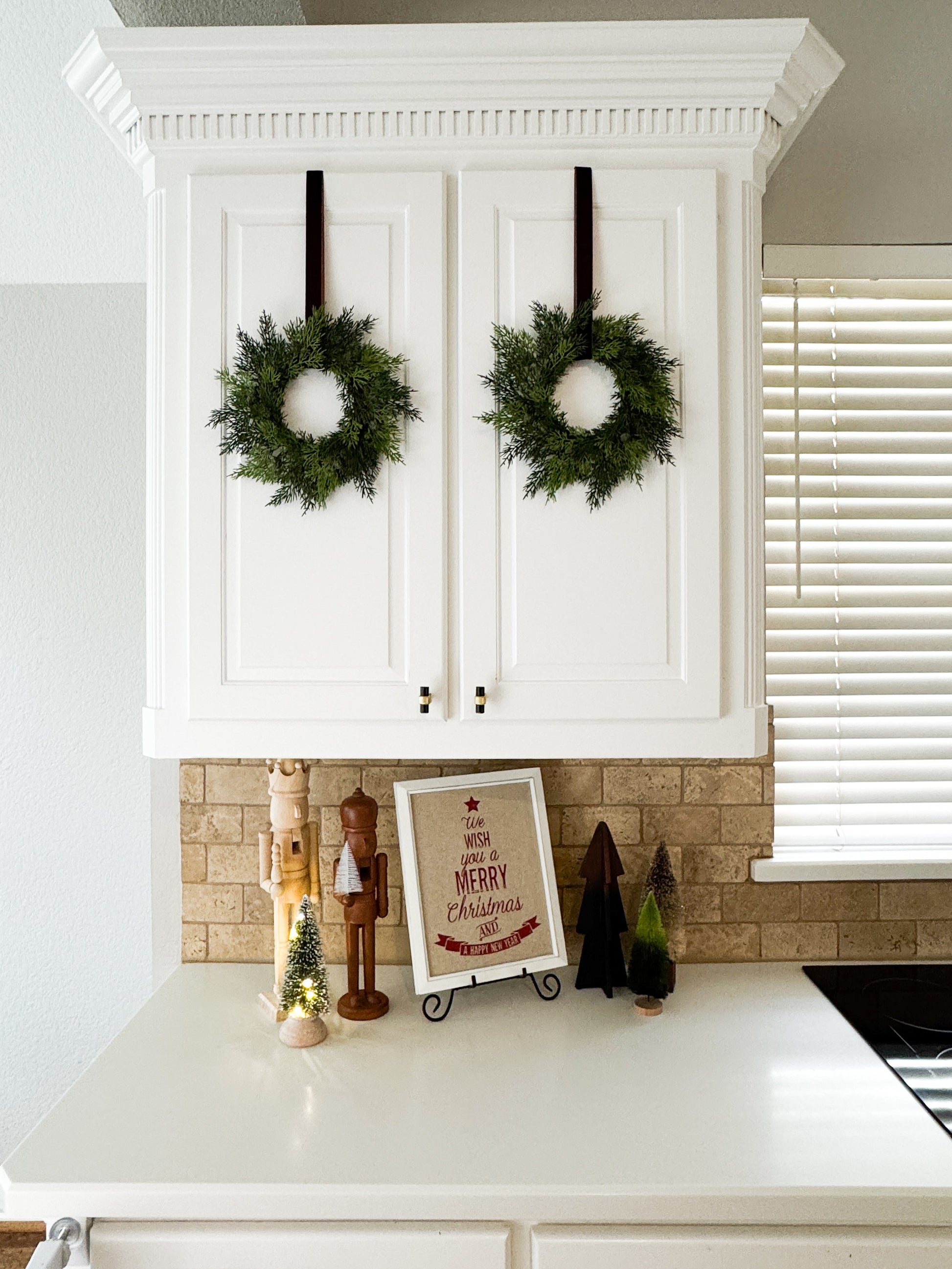 mini cedar wreaths hanging on a kitchen cabinet with Christmas-themed decor.