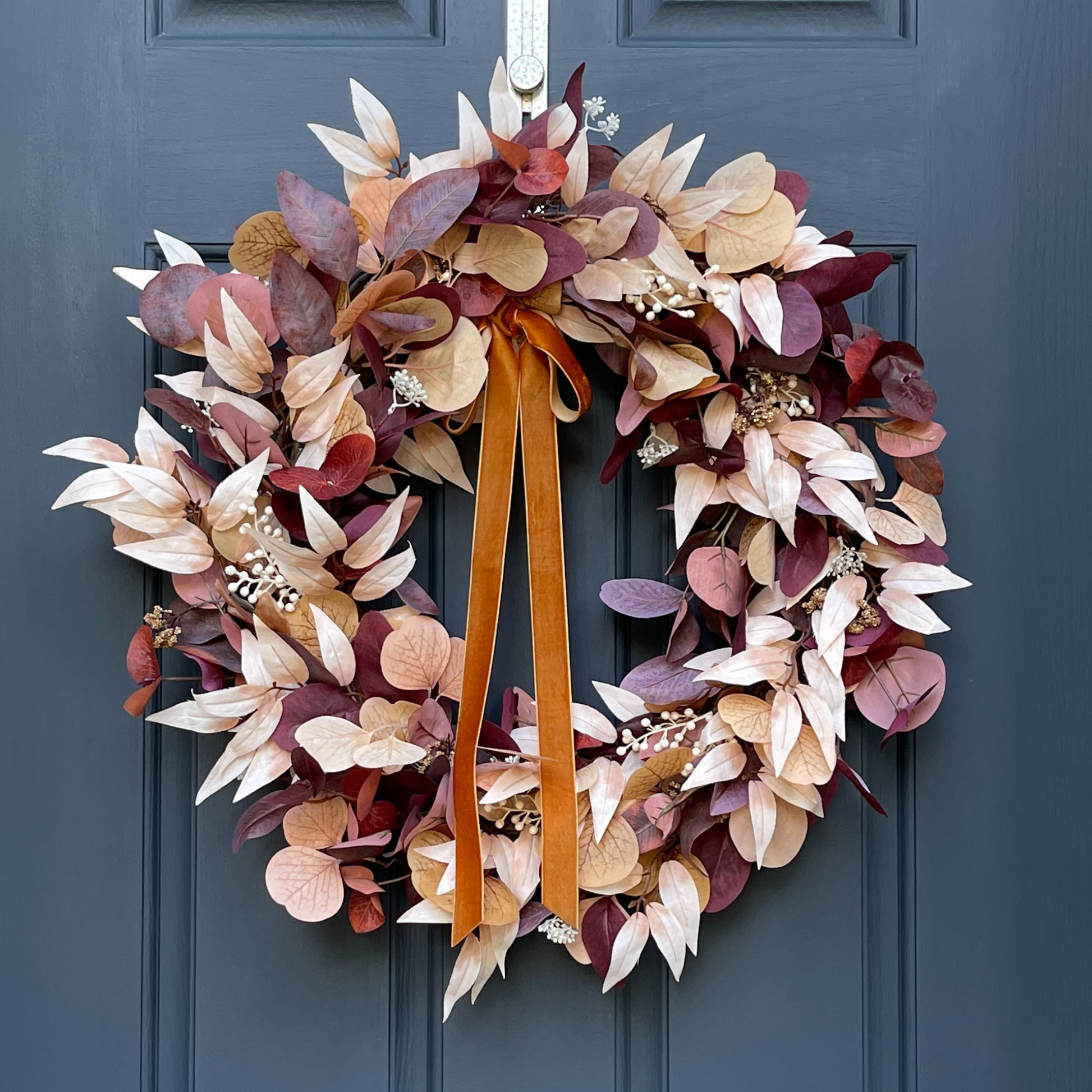 This neutral autumn wreath has been handcrafted on a grapevine base and is filled with various types of cream & rust eucalyptus and berries, and is finished with a velvet copper bow.  Wreath is hanging from a gold wreath hanger on a dark blue door.