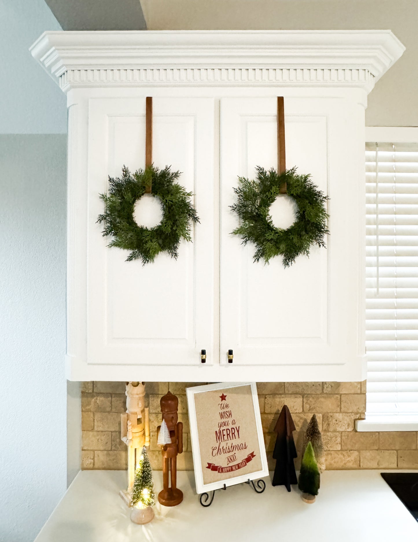 Mini cedar Christmas wreaths hanging on a kitchen cabinets with Christmas-themed decor.