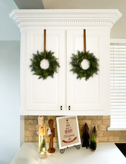 Mini cedar Christmas wreaths hanging on a kitchen cabinets with Christmas-themed decor.