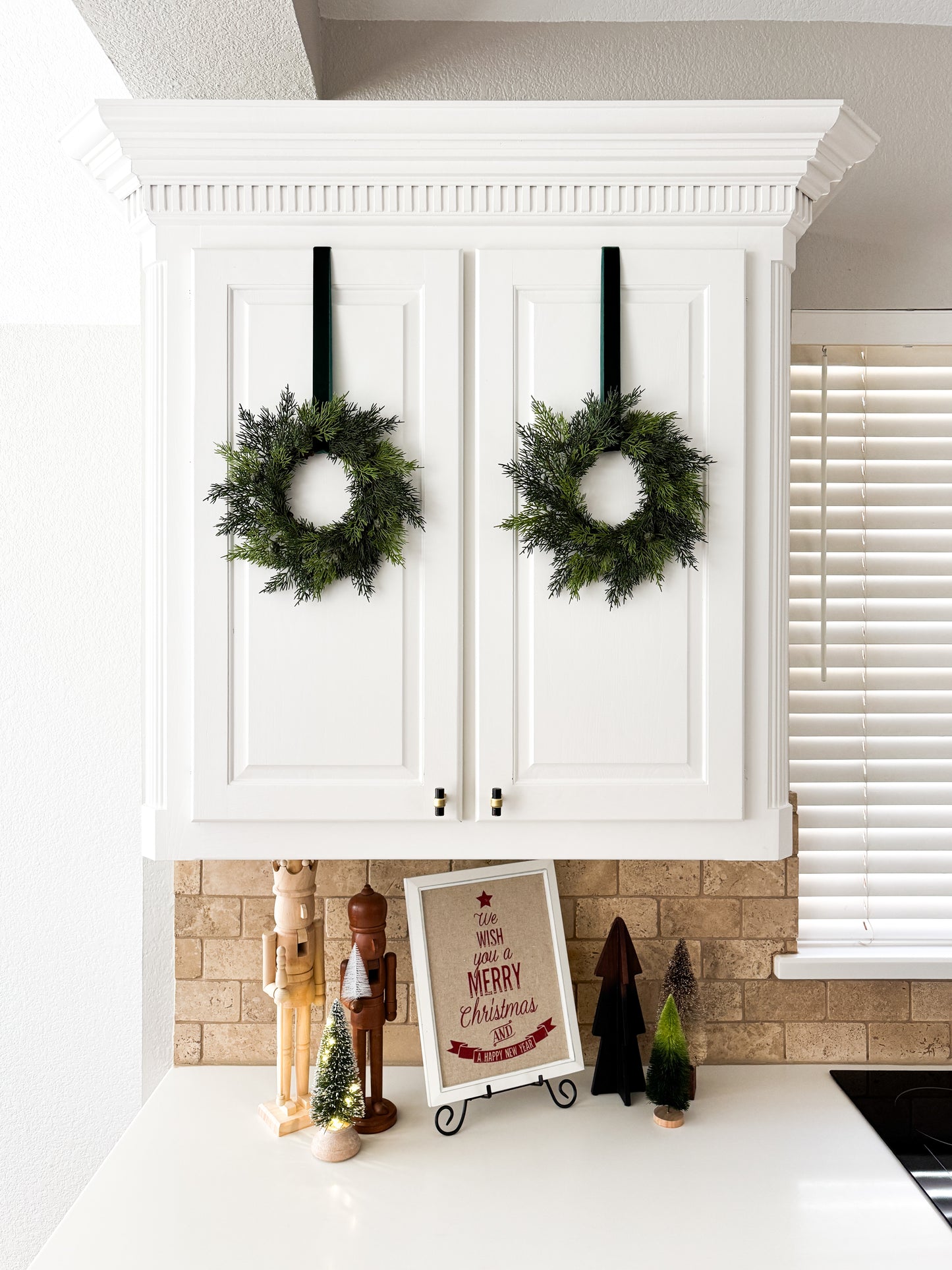 White kitchen cabinets with mini cedar holiday wreaths on a white countertop, Christmas decor.