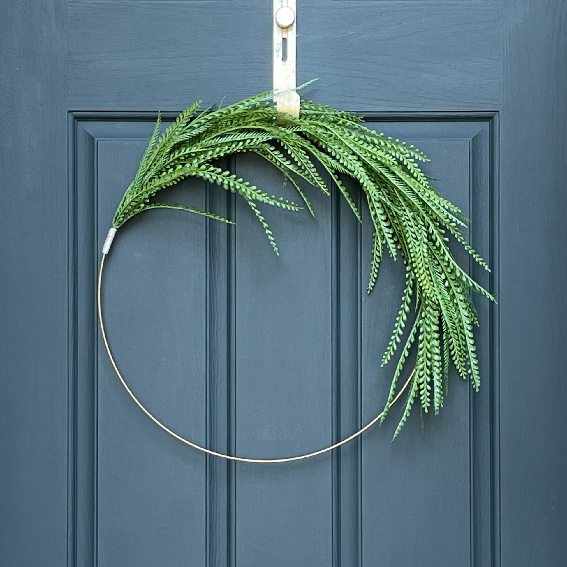 24" asymmetrical wreath, featuing green fern-like leaves, hanging from a gold wreath hanger on a dark blue door. 