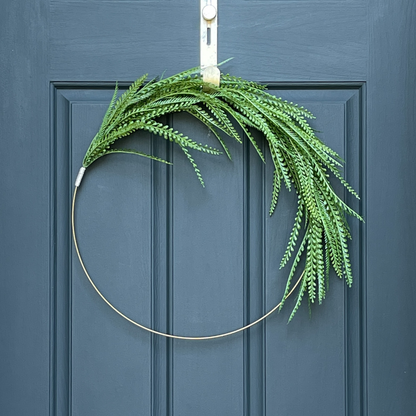 24" asymmetrical wreath, featuing green fern-like leaves, hanging from a gold wreath hanger on a dark blue door. 