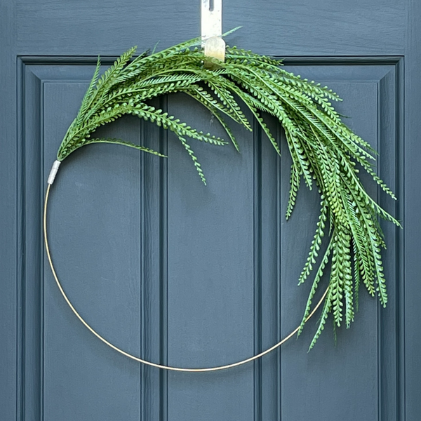 24" asymmetrical wreath, featuing green fern-like leaves, hanging from a gold wreath hanger on a dark blue door. 