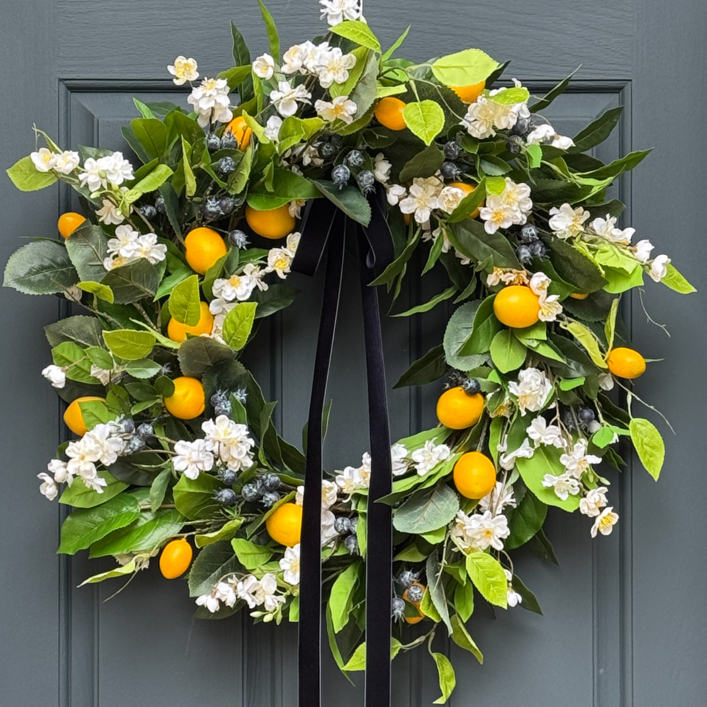 Decorative wreath with green leaves, white flowers, yellow fruits, and blueberries hanging on a door.
