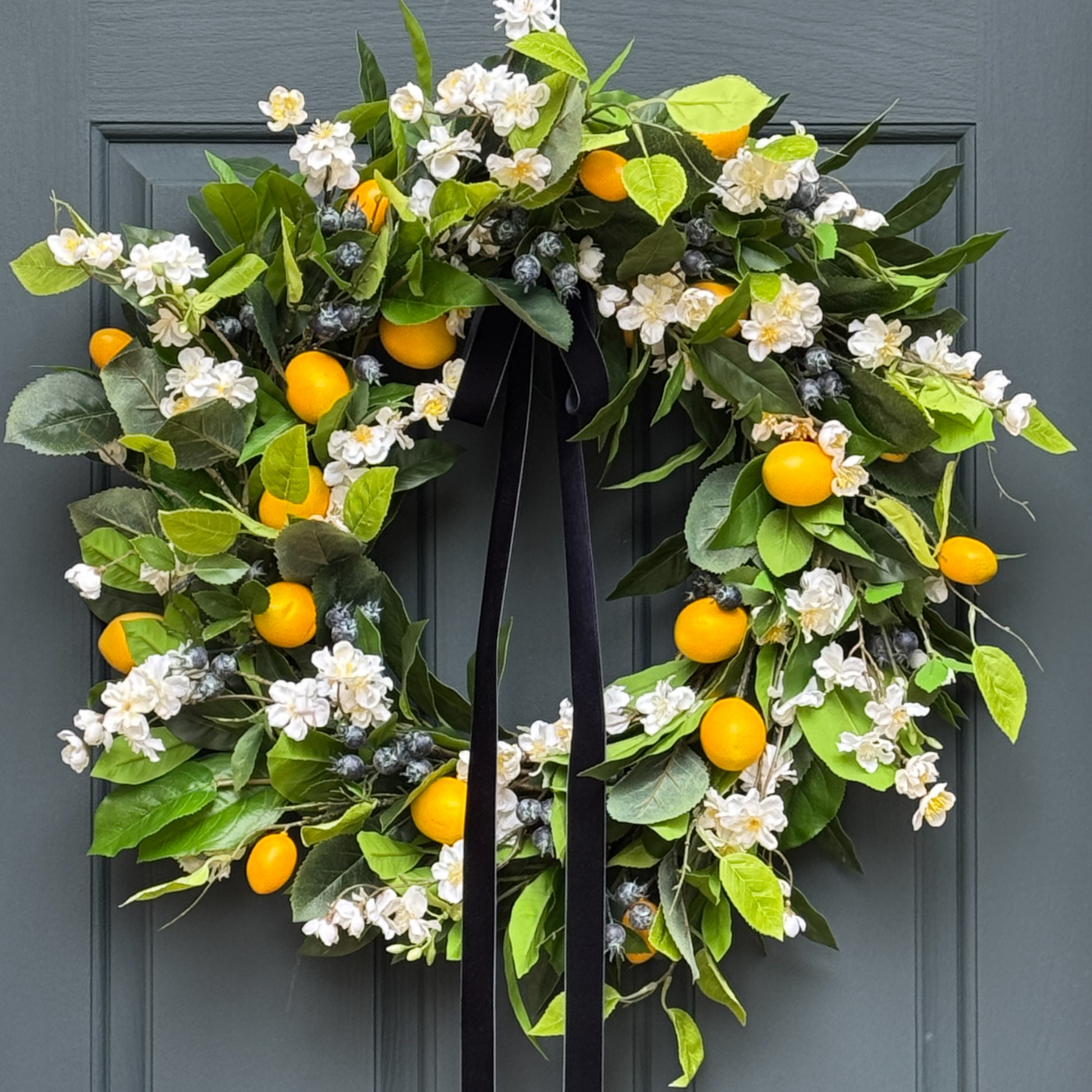 Decorative wreath with green leaves, white flowers, yellow fruits, and blueberries hanging on a door.