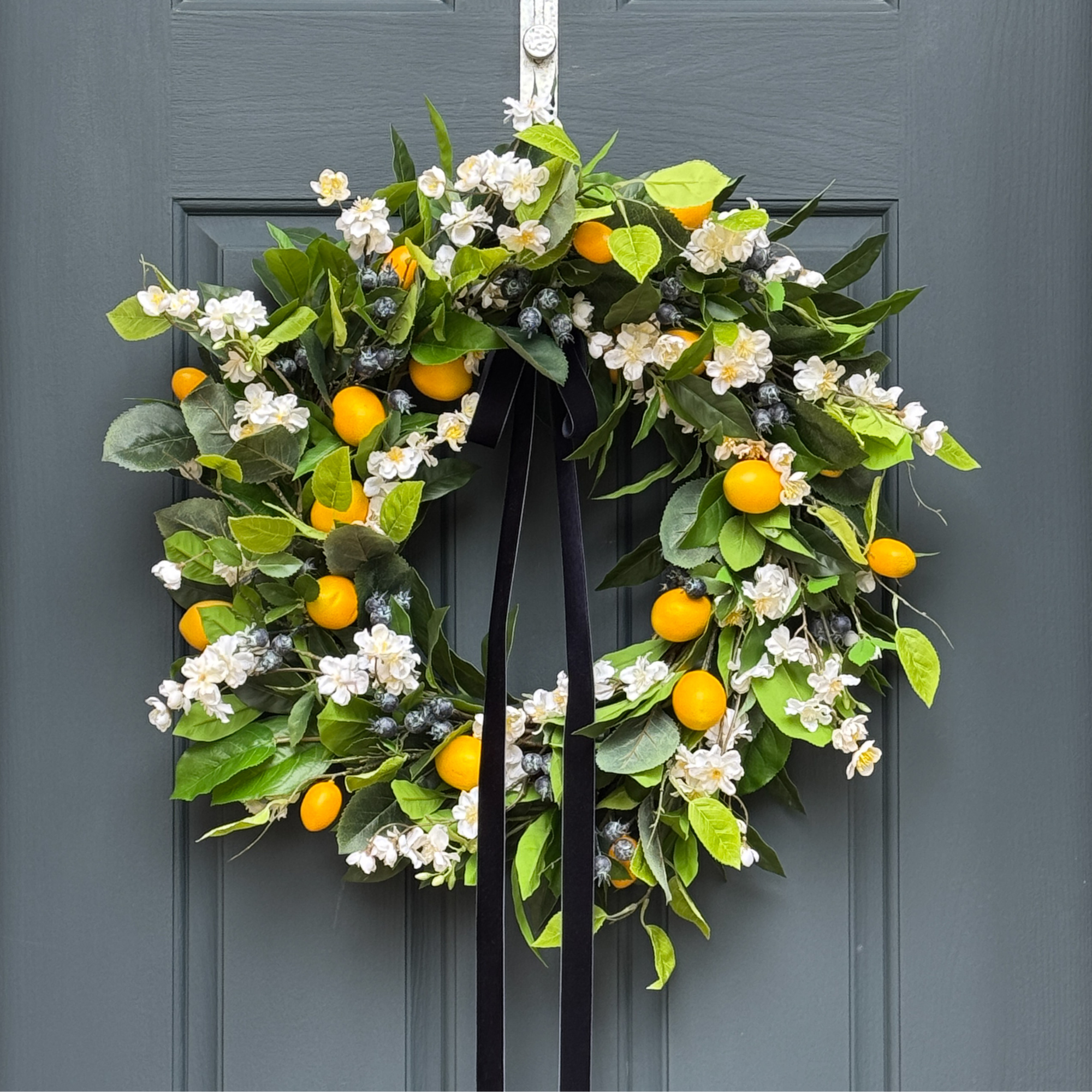 Decorative wreath with green leaves, white flowers, yellow fruits, and blueberries hanging on a door.