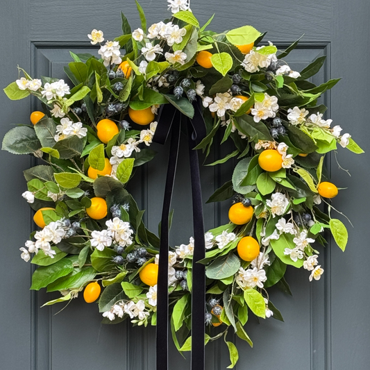 Decorative wreath with green leaves, white flowers, yellow fruits, and blueberries hanging on a door.