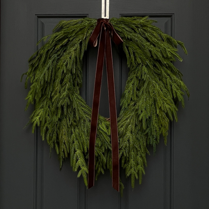 A faux Christmas greenery wreath with real touch norfolk pine and a dark brown velvet bow, hanging on a dark door.