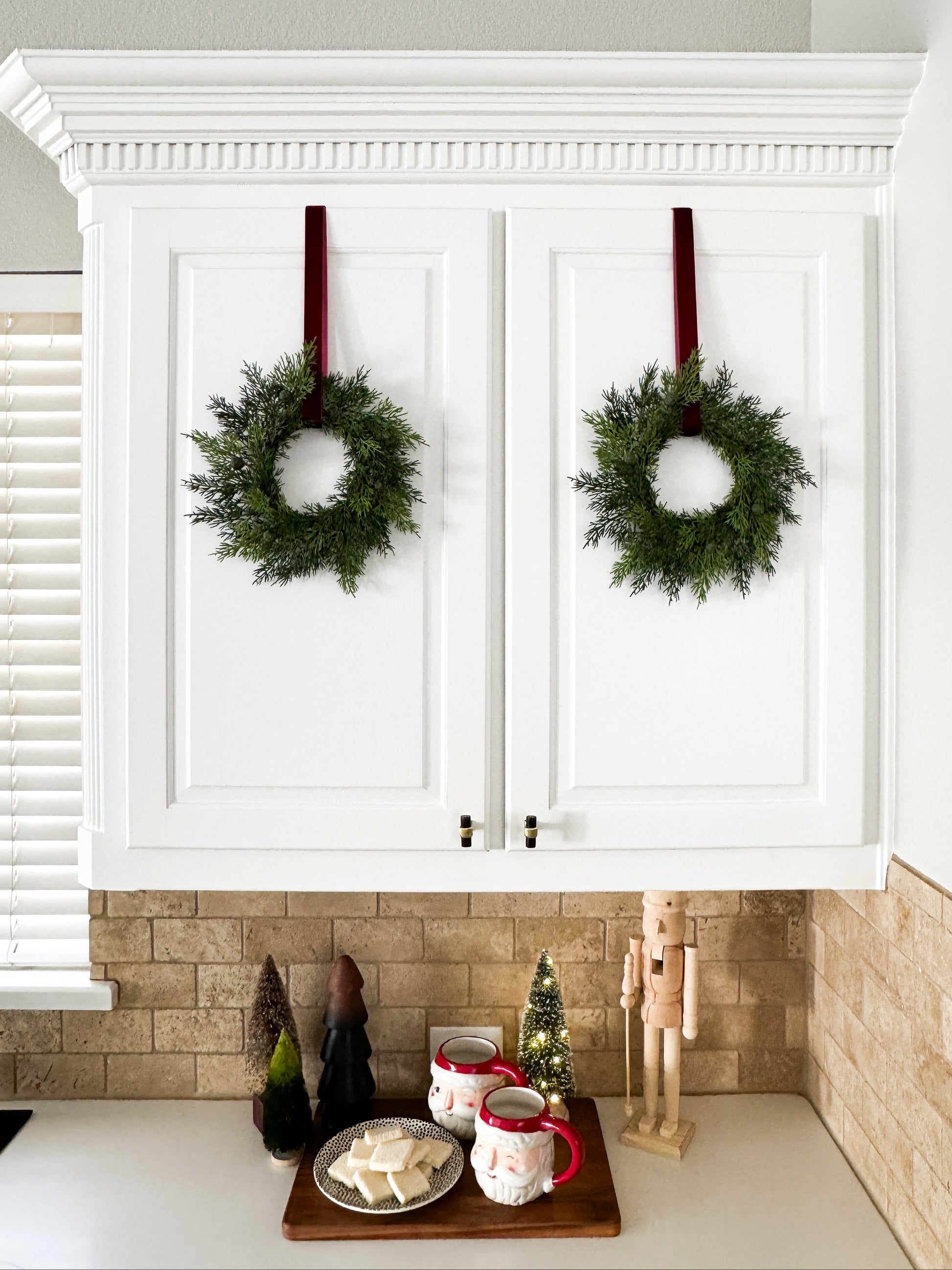 Two mini cedar Christmas wreaths hanging on white cabinets with a festive kitchen setting below.