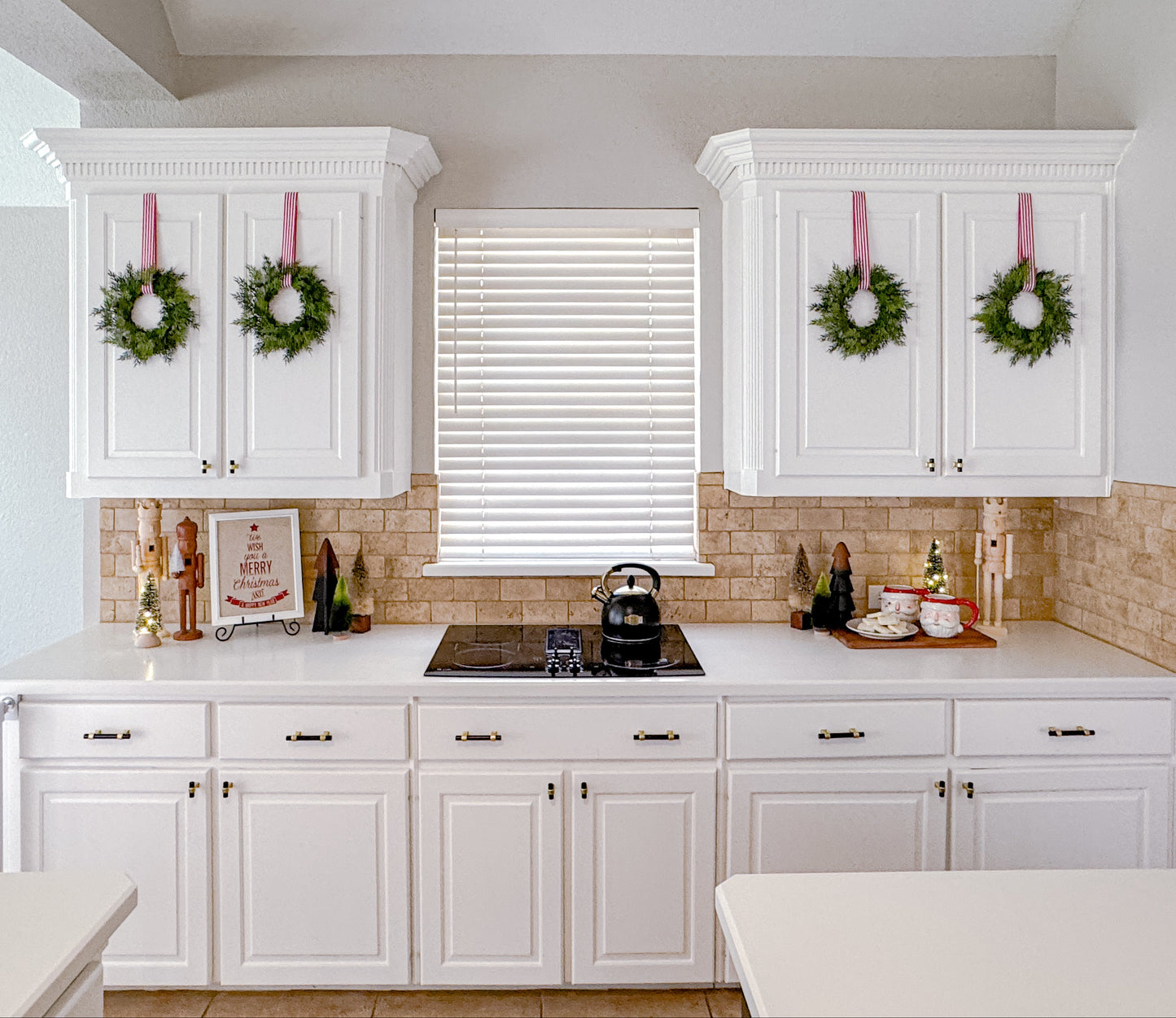 Kitchen with white cabinets, mini cedar Christmas wreaths, and a kettle on the stove.