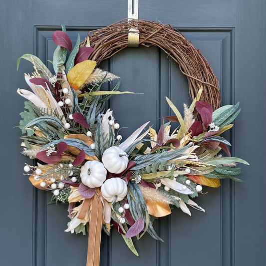 This rustic autumn wreath has been handcrafted on a grapevine base and is filled with eucalyptus, corn husks, sorghum sprays, and white berries, and is finished with a velvet antique gold bow. Wreath is hanging from a gold wreath hanger on a dark blue door.