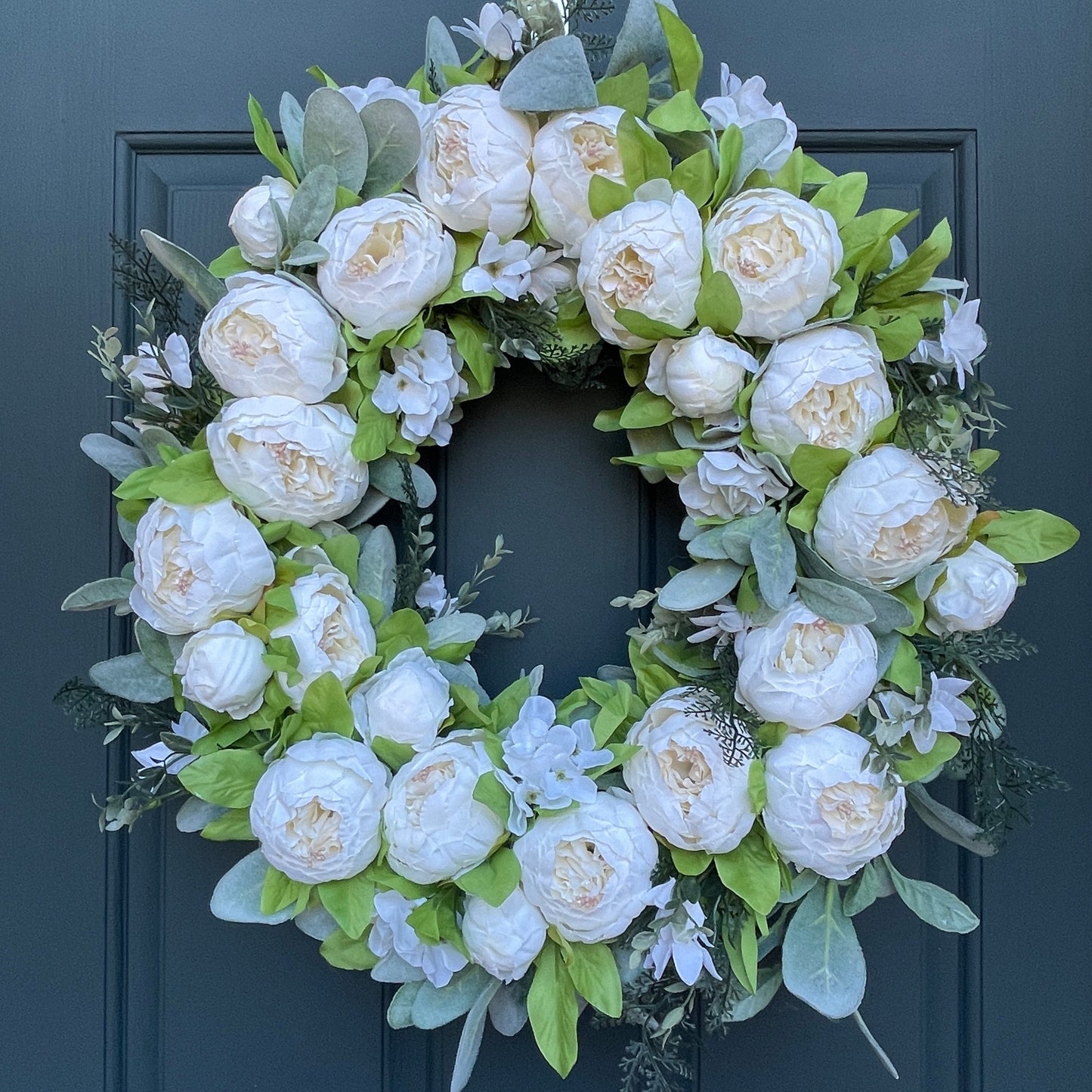 24" wreath has been handcrafted on a grapevine base and is trimmed with soft white peonies , hydrangeas, and other floral accents on a bed of flocked lambs ear. Wreath is hanging from a gold wreath hanger on a dark blue door.