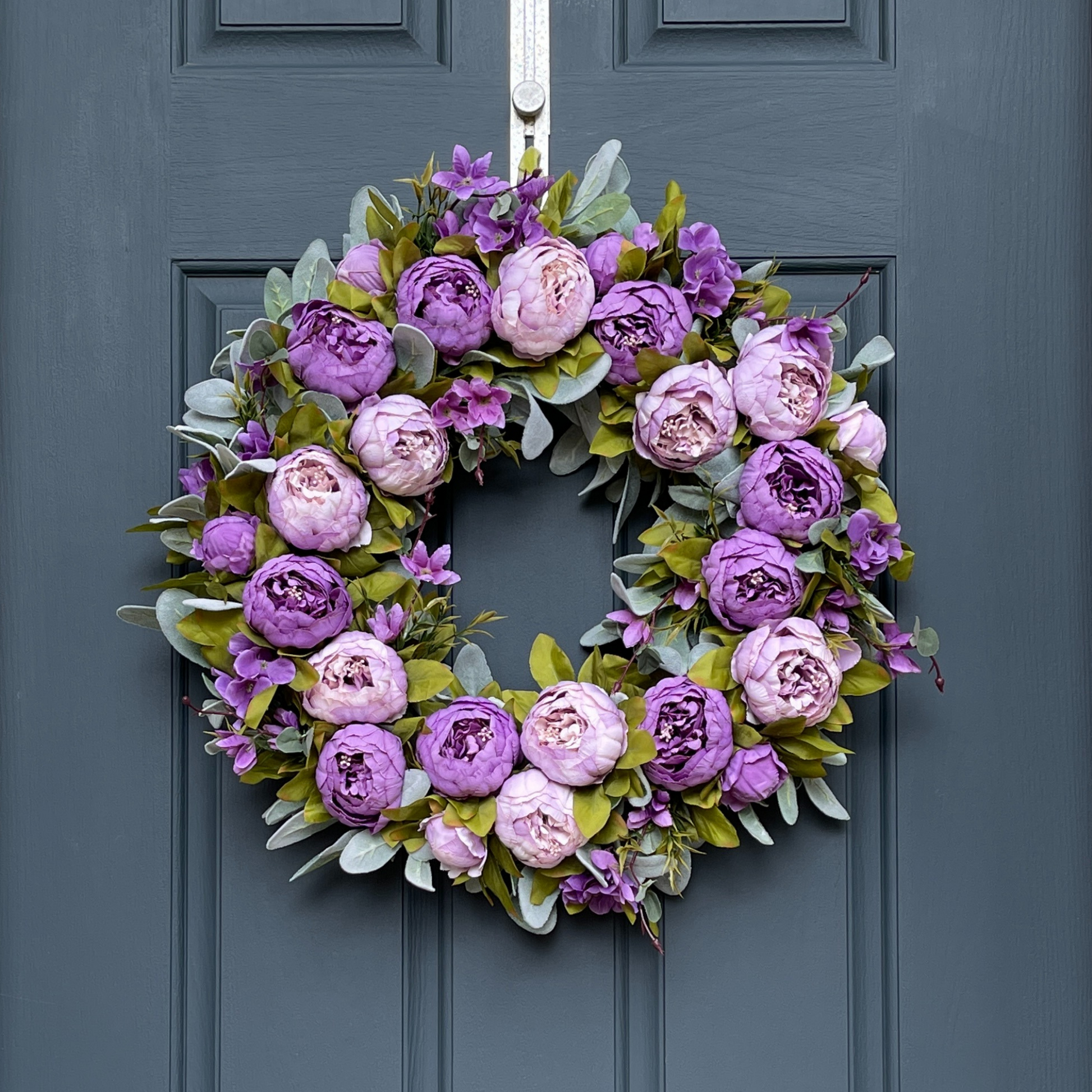 24" spring wreath. Wreath features two shades of purple peonies, peony buds, and hydrangeas, all on a bed of green flocked lambs ear. Wreath hangs from a gold hammered wreath hanger on a dark blue front door.