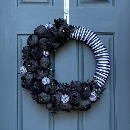 black and white halloween wreath made on a straw form that is wrapped with black and white striped ribbon with black peonies, leaves, black velvet pumpkins, and black and white striped pumpkins hanging from a gold wreath hanger on a dark blue door