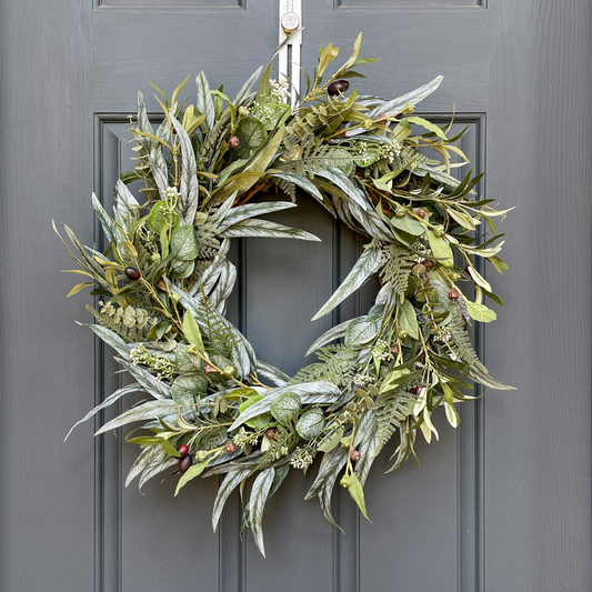 24" every-day use greenery wreath. The wreath is filled with various types of eucalytpus, fern leaves, and olive branches. Wreath hangs from a hammered gold wreath hanger on a dark blue front door.