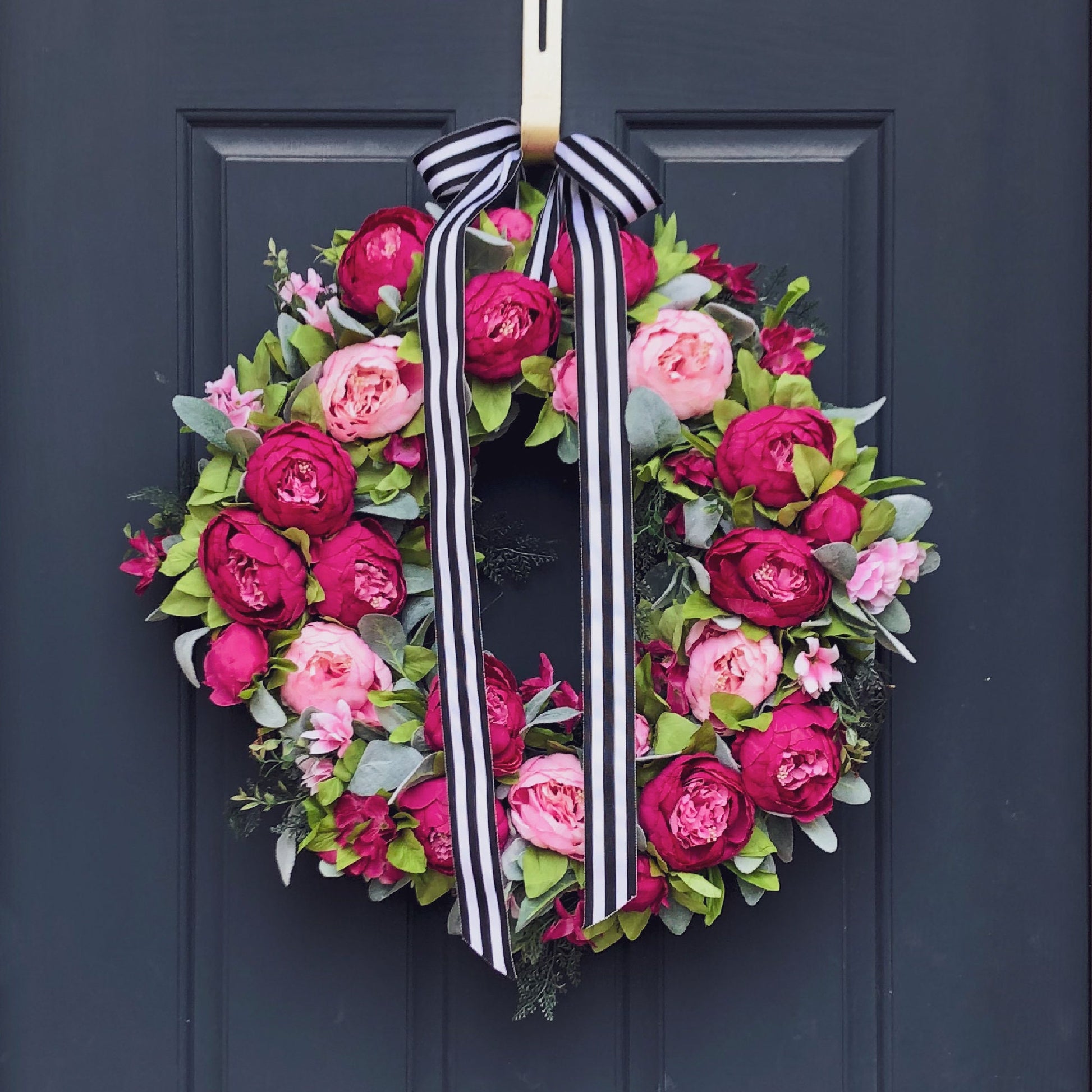 24" wreath has been handcrafted on a grapevine base and is trimmed with peonies in two shades of pink (pale pink & hot pink), hydrangeas, and other floral accents on a bed of flocked lambs ear and is finished with a black & white striped bow. 