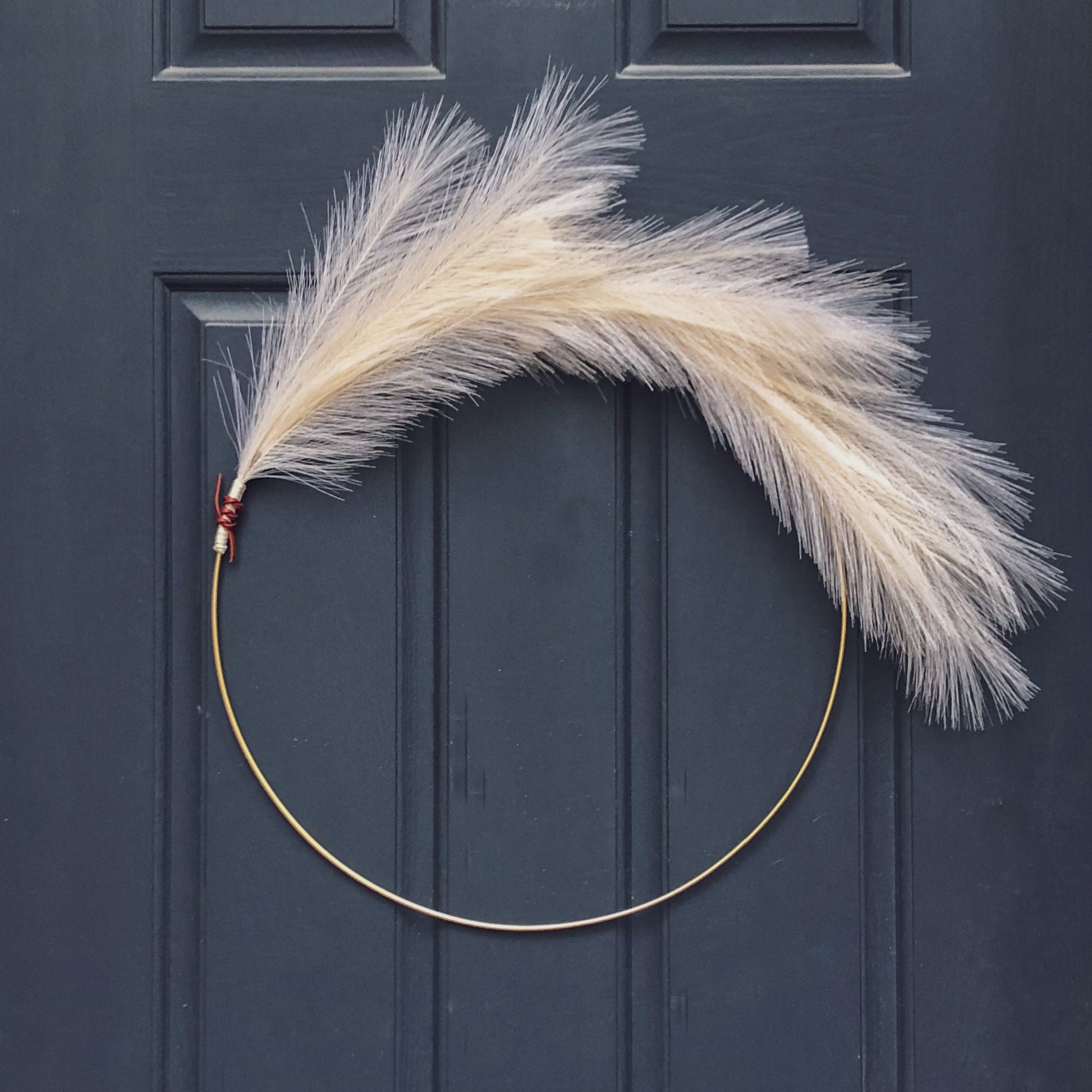 Asymmetrical 24" matte gold metal hoop with artificial cream pampas grass sprays. One end of pampas grass is wrapped with white cotton twine and shoelace leather. Wreath is hanging on a dark blue front door.