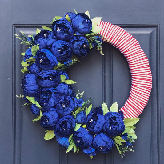 24" straw wreath form wrapped with red and white striped ribbon. Half of the wreath is covered with navy blue peony blooms and buds with green peony leaves. Wreath is hanging on a dark blue front door.