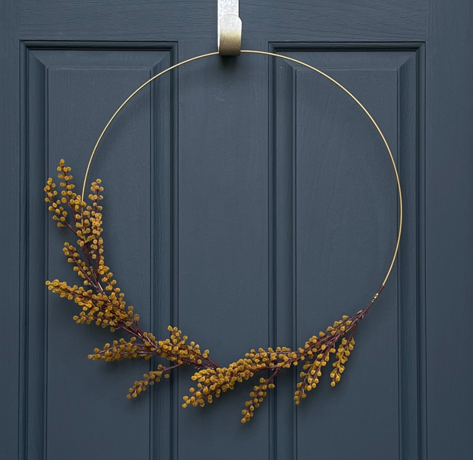 20" fall wreath. Gold hoop with greenish-yellow mimosa sprays. Stem of mimosa spray is wrapped with gold floral wire. Wreath is hanging from a gold hammered wreath hanger on a dark blue front door.