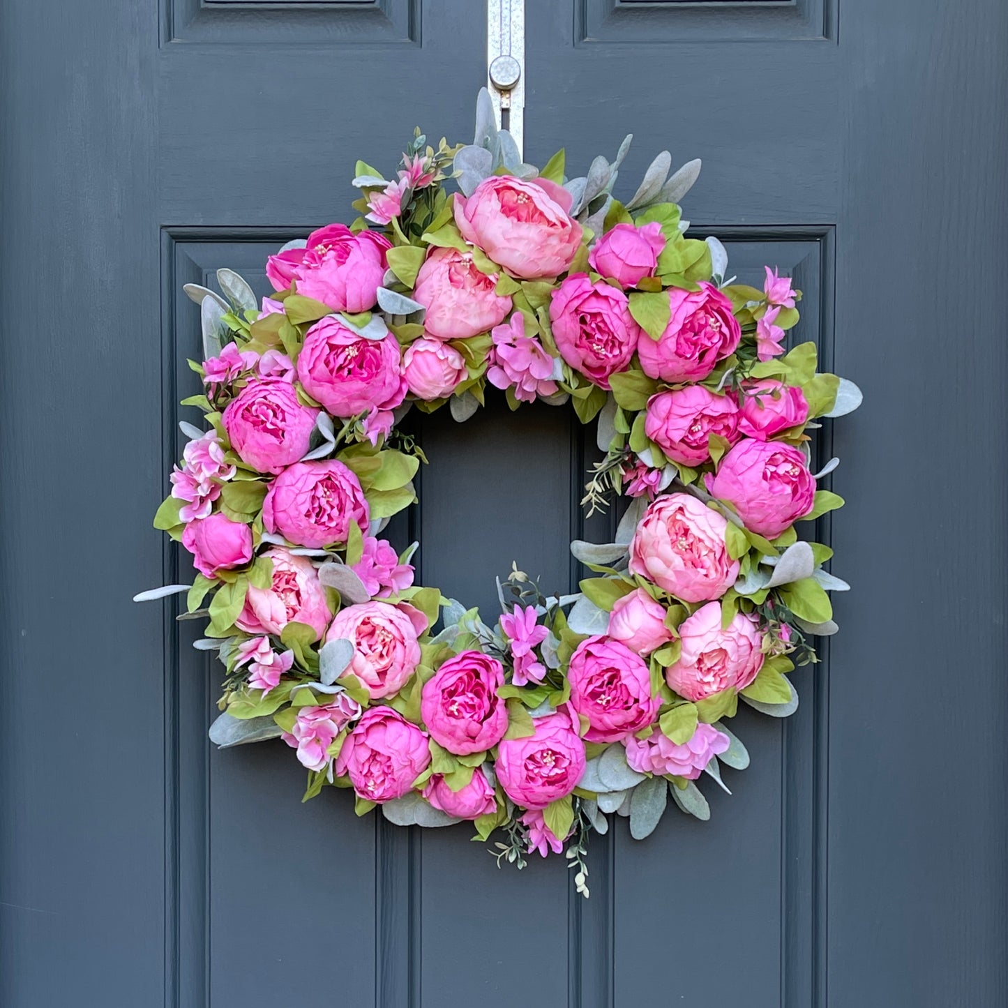 24" wreath has been handcrafted on a grapevine base and is trimmed with peonies in two shades of pink peonies (pale pink & bright pink), hydrangeas, and other floral accents on a bed of flocked lambs ear.