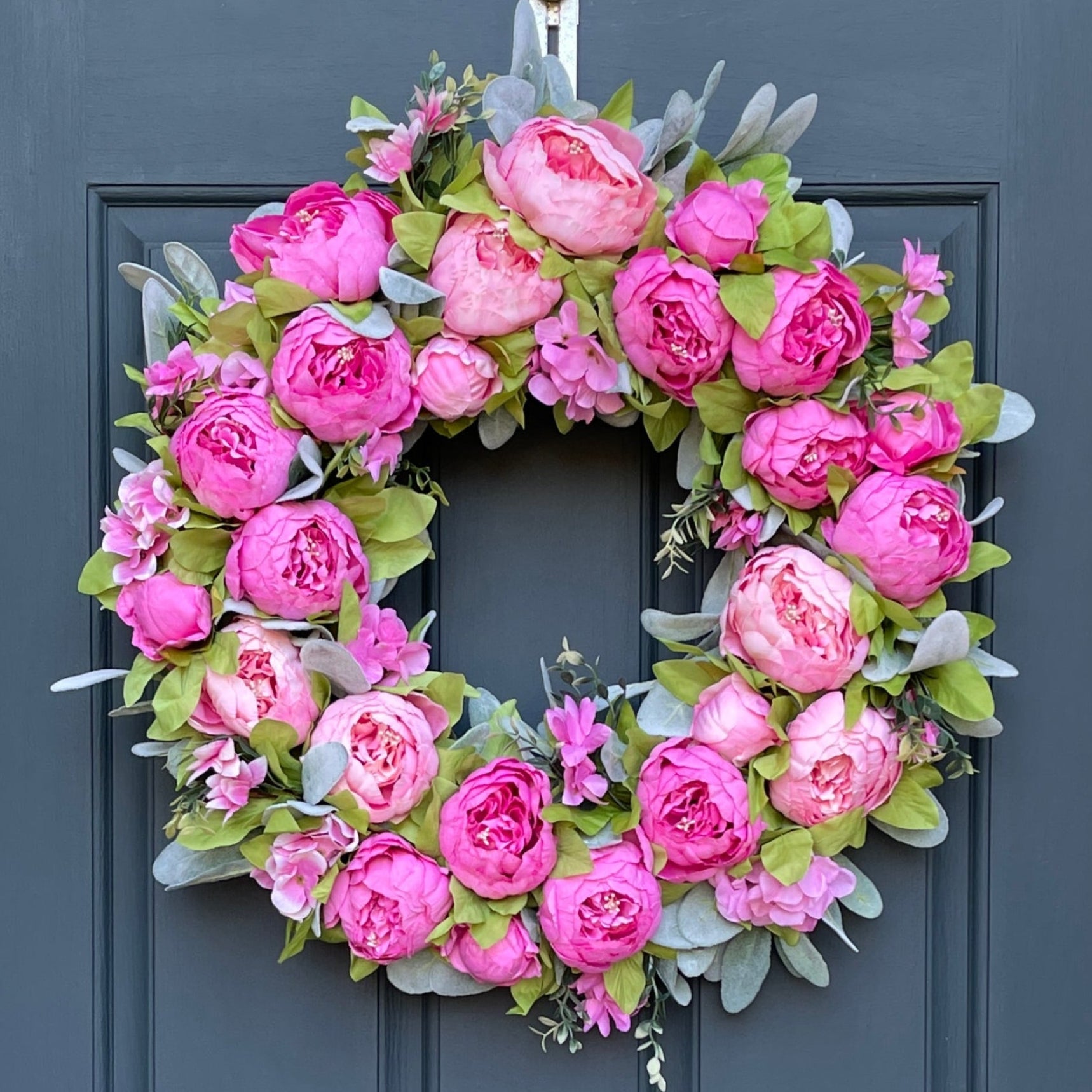 24" wreath has been handcrafted on a grapevine base and is trimmed with peonies in two shades of pink peonies (pale pink & bright pink), hydrangeas, and other floral accents on a bed of flocked lambs ear.