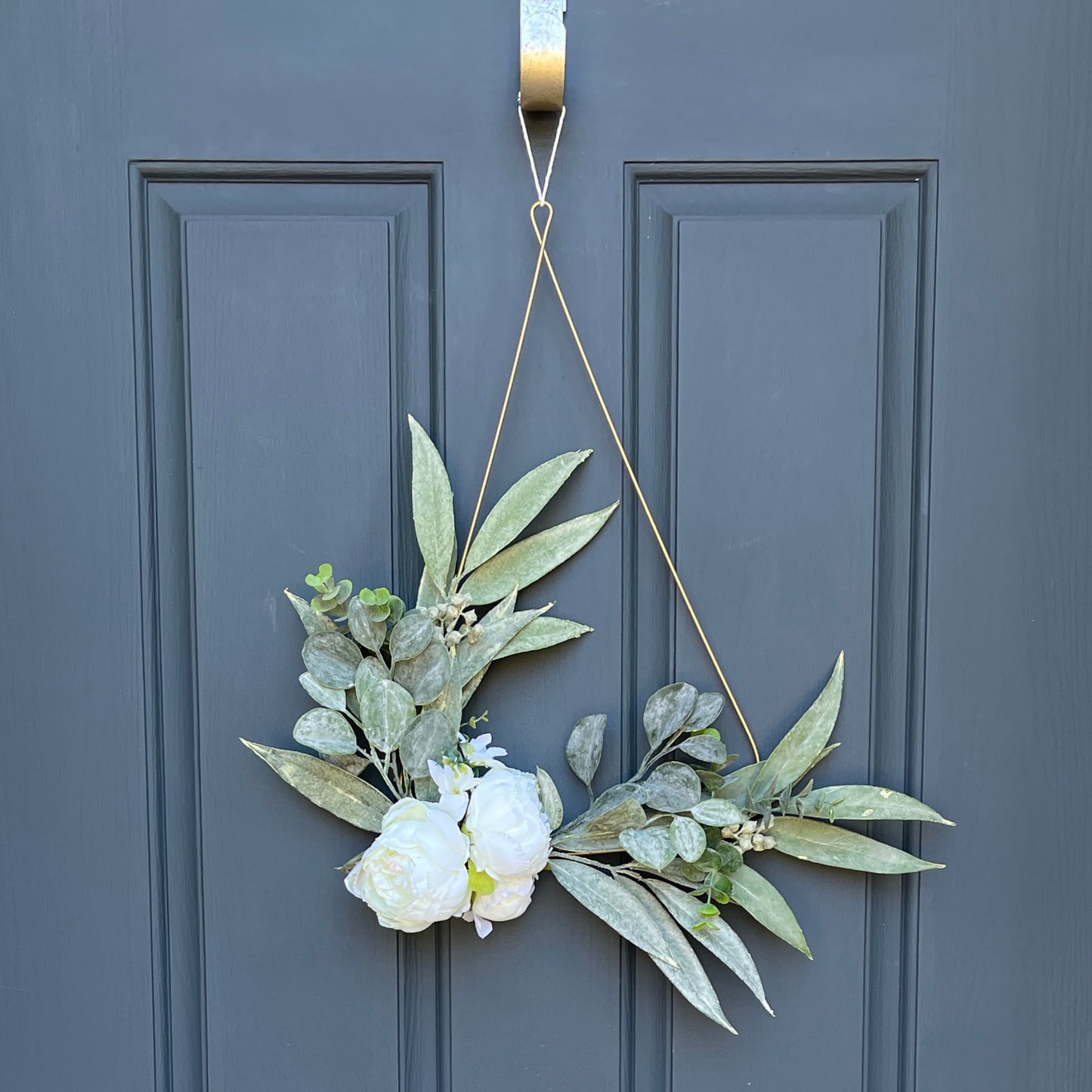 Matte gold, tear-drop shaped metal wreath base with eucalyptus greenery, boxwood greenery, and two white peony blooms and one white peony bud. Wreath has a cotton twine hanger. Wreath is hanging from a gold wreath hanger on a dark blue front door.