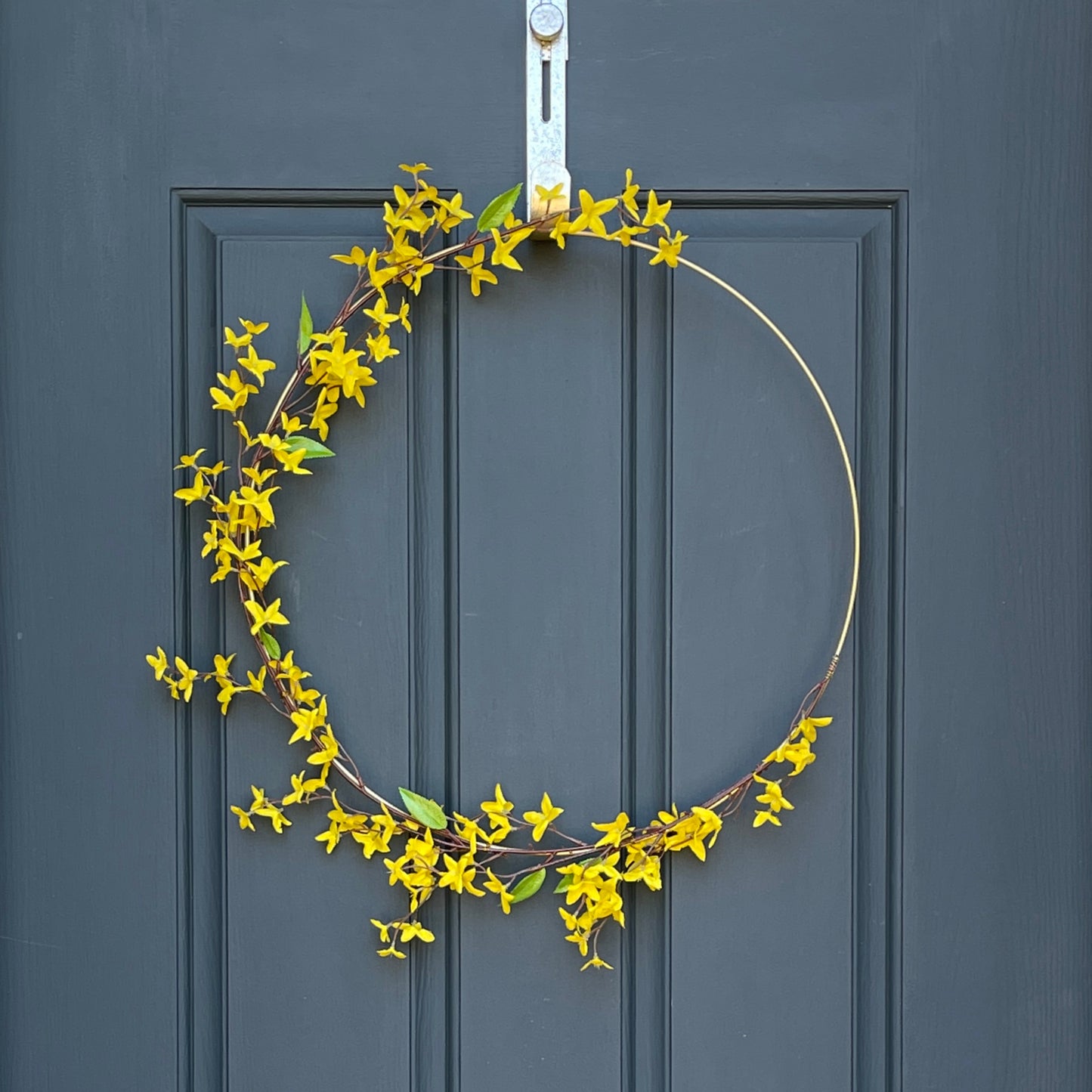 24" matte gold metal hoop with branches of yellow forsythia stems. Stems are attached with gold floral wire. Wreath is hanging from a gold wreath hanger on a dark navy front door.