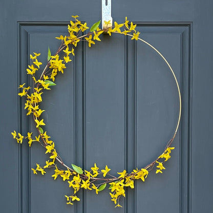 24" matte gold metal hoop with branches of yellow forsythia stems. Stems are attached with gold floral wire. Wreath is hanging from a gold wreath hanger on a dark navy front door.